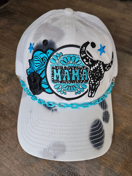 Tie dye trucker hat in black and turquoise with "mama", bullhead, and roses patches. Accented with a turquoise hat chain. snap back closure for adjustment.