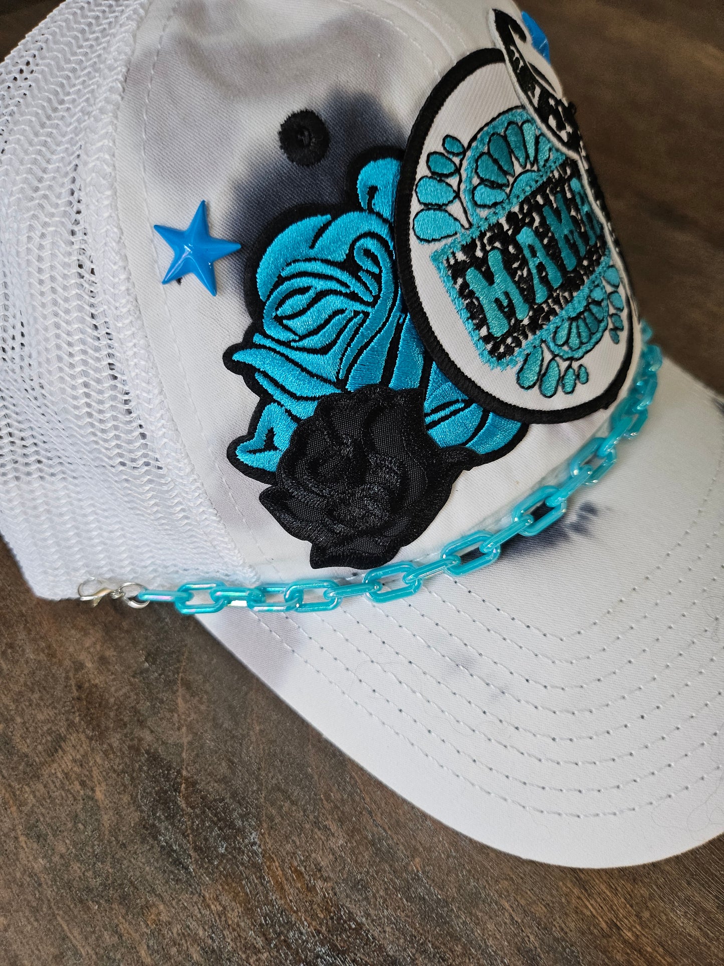 Tie dye trucker hat in black and turquoise with "mama", bullhead, and roses patches. Accented with a turquoise hat chain. snap back closure for adjustment.